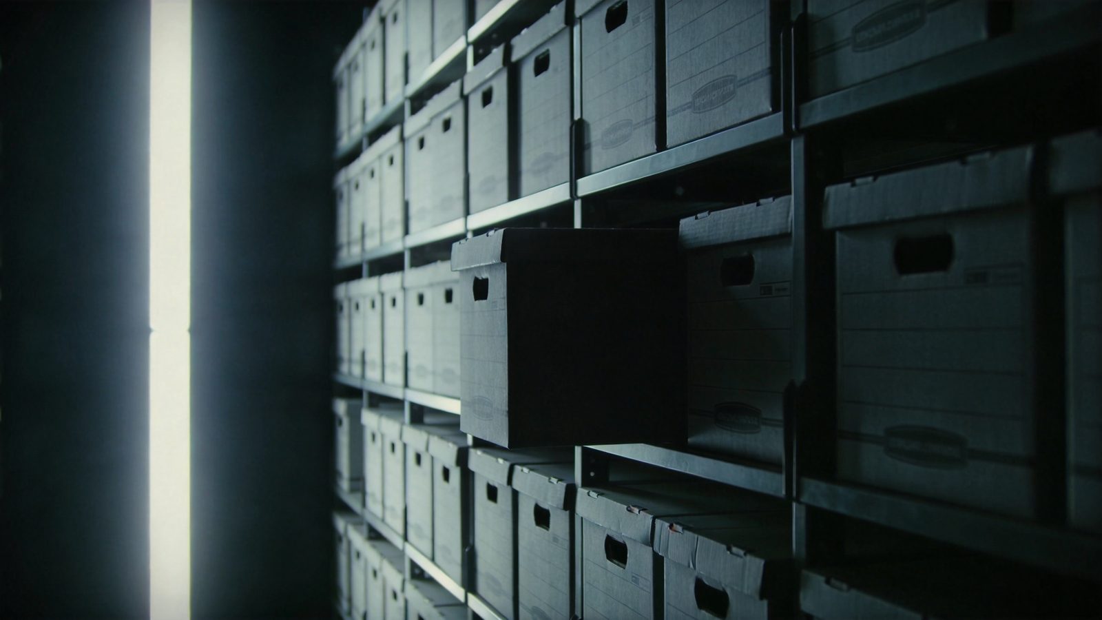 Atmospheric image: dark archive shelves full of anonymous file boxes, a cold shaft of light from the left, one box pulled halfway out — symbolic of sealed records and the fight over disclosure.